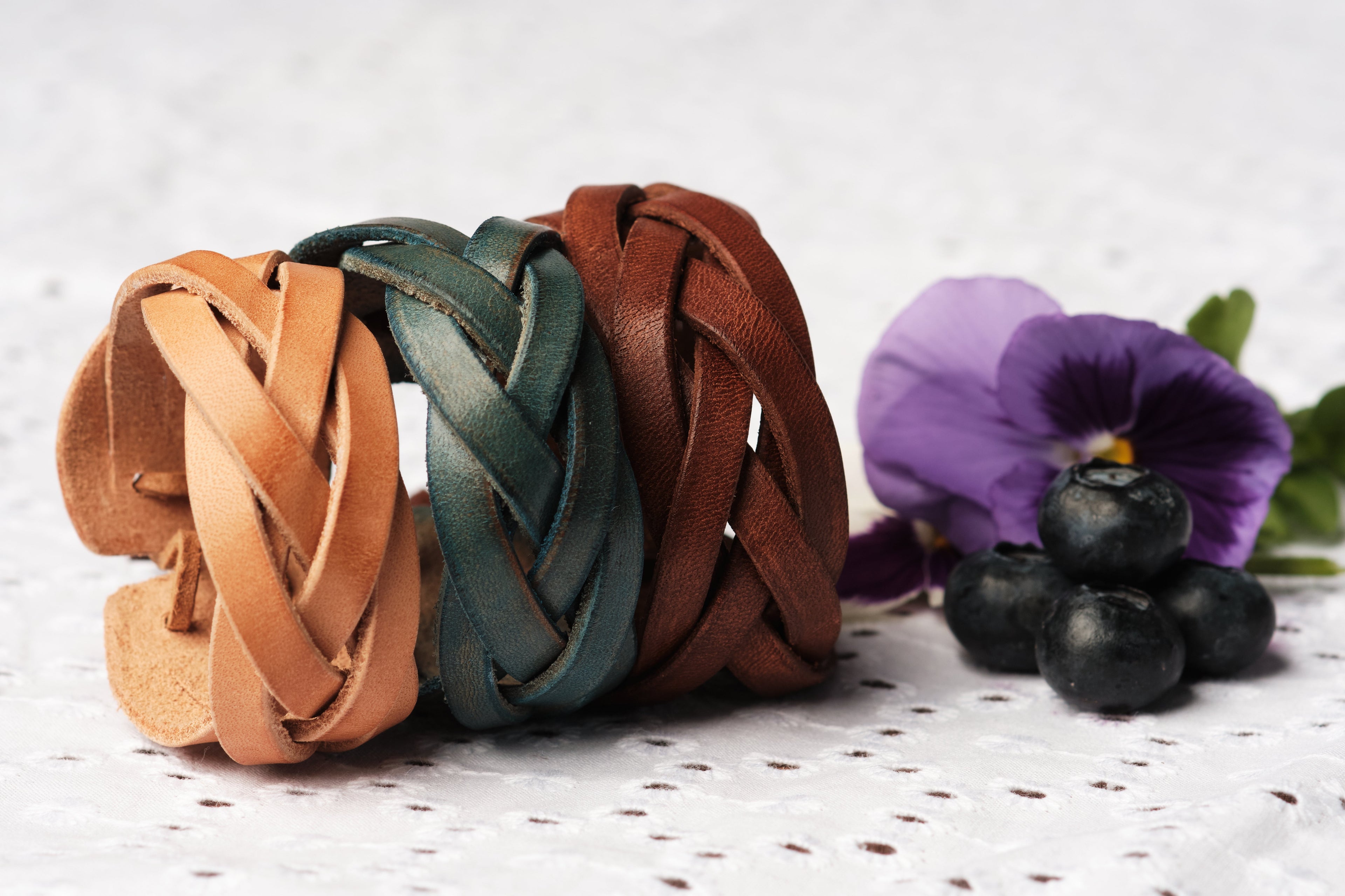 Three braided leather bracelets in brown, green, and red on a textured surface with a purple flower and blueberries.