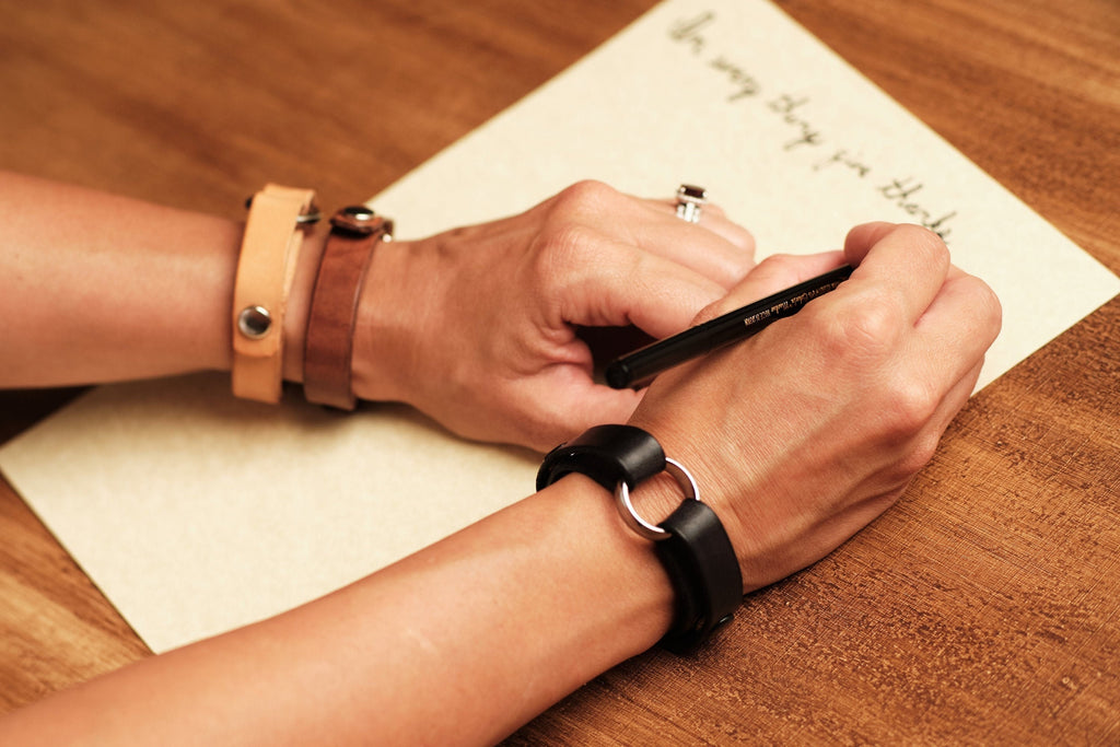 Person writing on a piece of paper with a pen, wearing a watch and bracelet on a wooden surface.