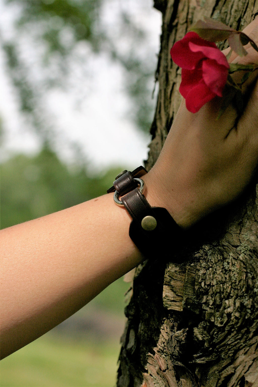 Person wearing a black leather bracelet with a red flower against a tree