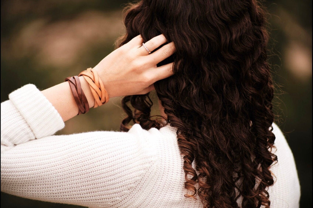 Person with long, wavy hair touching it, wearing a white sweater and multiple bracelets.