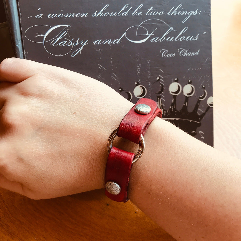 Red leather bracelet with silver accents on a wrist, with a book in the background featuring text.