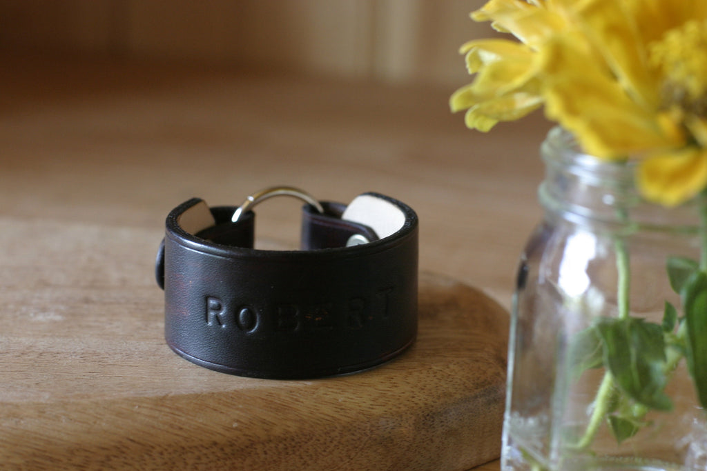 Black leather wear the ring bracelet with 'Rovcom' branding on a wooden surface next to a jar of yellow flowers.